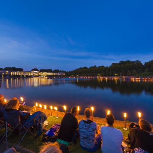 Menschen sitzen mit Picknickdecken um den Hollersee und lauschen einem klassischen Konzert 
